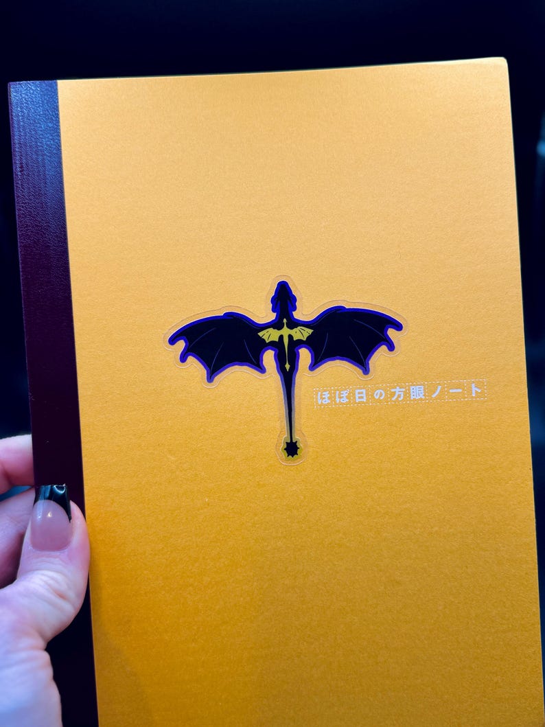 May include: A mustard-yellow notebook with a dark blue and gold dragon design. The dragon has outstretched wings and a long tail. Japanese text is printed on the upper right corner of the notebook.