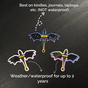 May include: Three bat-shaped stickers with iridescent wings and yellow and blue accents. The stickers are weather/waterproof for up to 2 years and are best on kindles, journals, and laptops. The text on the image says "Best on kindles, journals, laptops, etc. (NOT waterproof)" and "Weather/waterproof for up to 2 years."
