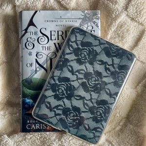 May include: A hardcover book titled "The Serpent & the Wings of Night" with a green and white cover, next to a clear phone case with a black floral lace pattern. The items rest on a cream-colored, textured surface.