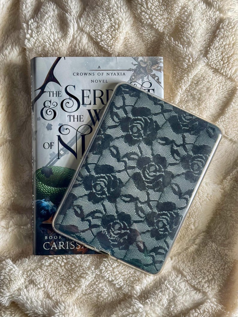 May include: A hardcover book titled "The Serpent & the Wings of Night" with a green and white cover, next to a clear phone case with a black floral lace pattern. The items rest on a cream-colored, textured surface.