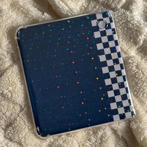 May include: A clear tablet case with a navy blue background covered in colorful star-shaped glitter. The right side of the case features a black and white checkered pattern. The case is designed to protect a tablet.