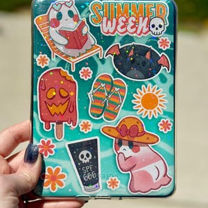 May include: A clear phone case decorated with summer-themed stickers. The stickers include a ghost reading a book, a bat, a popsicle, flip-flops, a sun, and a tube of SPF 666 sunscreen. The words "SUMMER WEEK" are also visible.