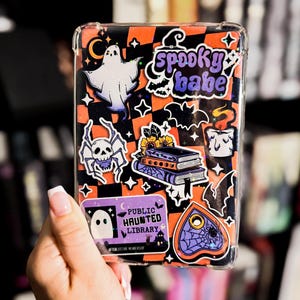 Könnte beinhalten: Eine transparente Handyhülle, verziert mit Halloween-Stickern. Die Aufkleber zeigen einen Geist, eine Spinne mit Totenkopf, Bücher, eine Kerze, eine Fledermaus und die Worte "spooky babe". Der Hintergrund ist orange und schwarz.
