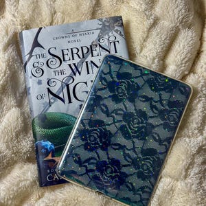 May include: A hardcover book titled "The Serpent & the Wings of Night" with a green snake illustration, next to a phone case with a dark blue floral lace pattern. Both items rest on a fluffy, cream-colored blanket.