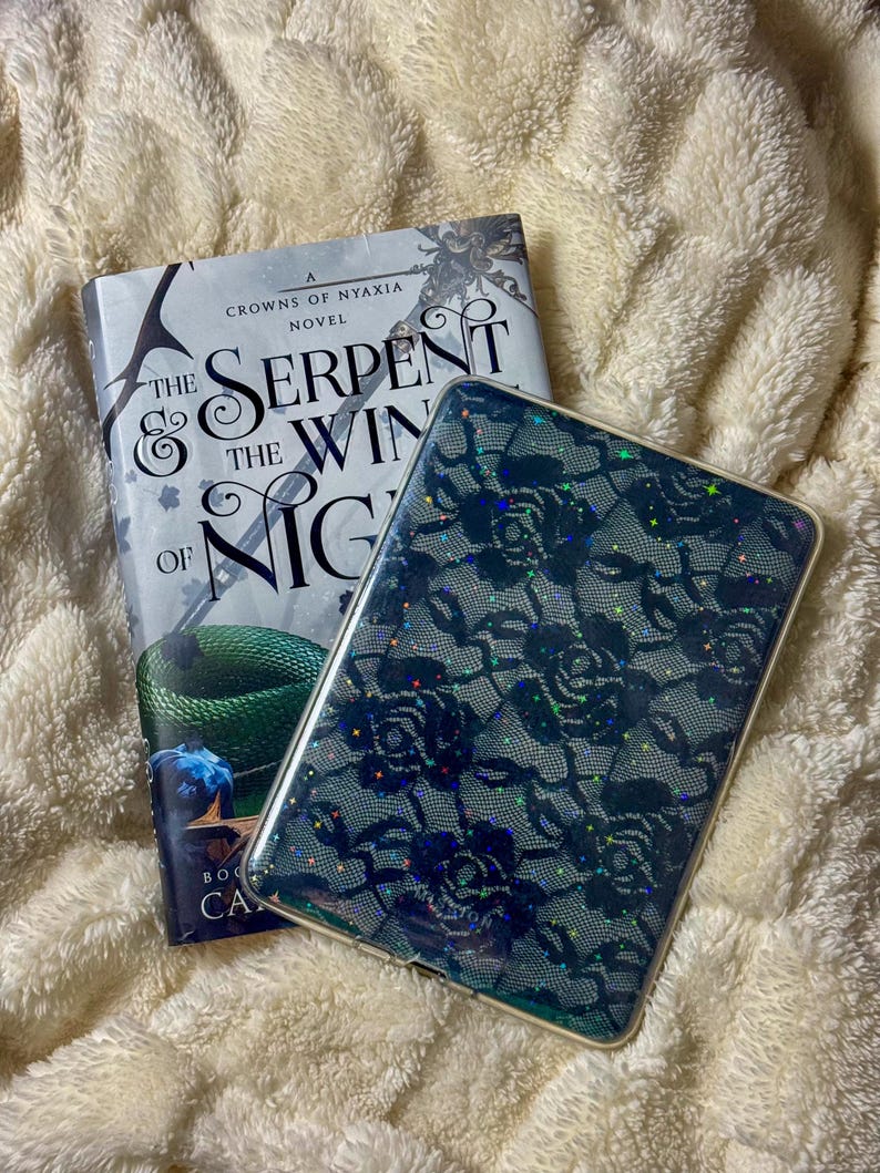 May include: A hardcover book titled "The Serpent & the Wings of Night" with a green snake illustration, next to a phone case with a dark blue floral lace pattern. Both items rest on a fluffy, cream-colored blanket.
