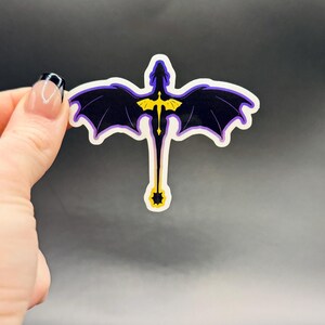 May include: A sticker featuring a stylized dragon with outstretched wings. The dragon is primarily black with purple wing edges and a yellow emblem on its chest. The sticker has a white border and is held against a dark background.