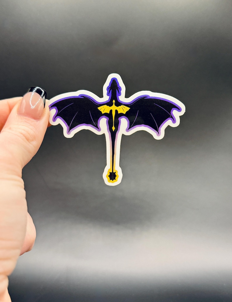 May include: A sticker featuring a stylized dragon with outstretched wings. The dragon is primarily black with purple wing edges and a yellow emblem on its chest. The sticker has a white border and is held against a dark background.