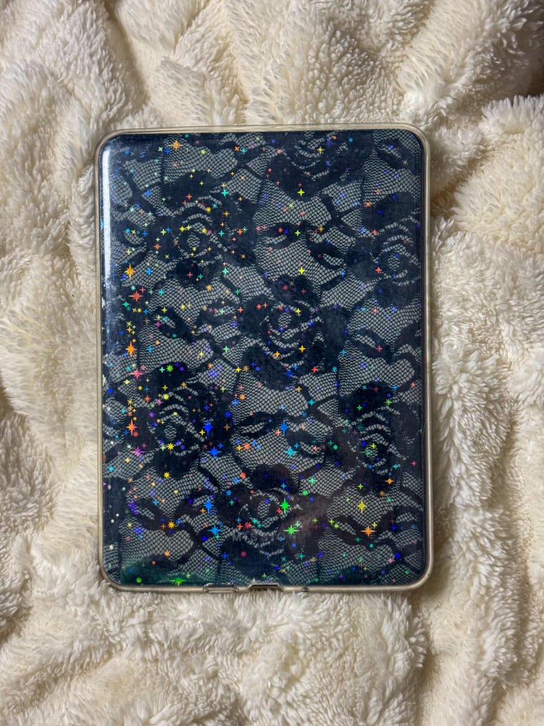 May include: A rectangular phone case with a black lace pattern and iridescent glitter. The case has a clear border and is set against a white, fluffy background.