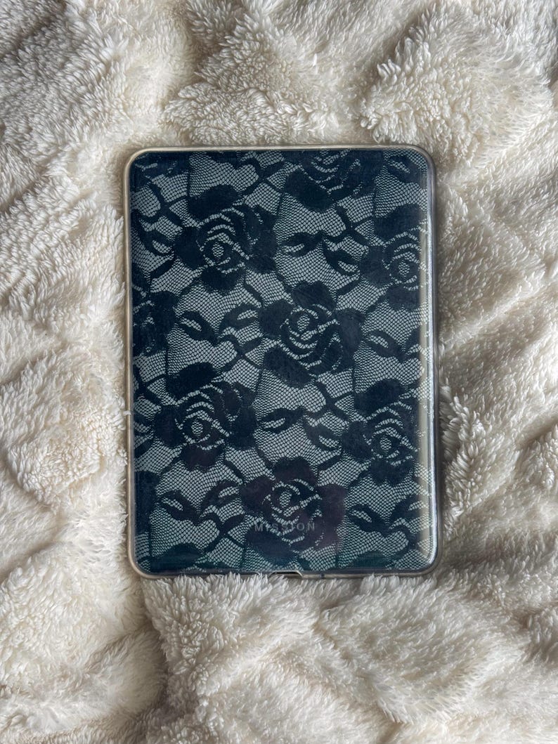 May include: A rectangular, clear phone case featuring a black lace rose pattern. The case has a transparent border and is set against a fluffy, white background. The lace design is detailed and covers the entire back of the case.