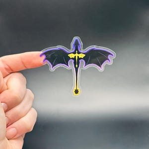 May include: A black dragon sticker with purple and yellow accents. The dragon has outstretched wings and a long tail. The sticker is held between two fingers against a dark background.