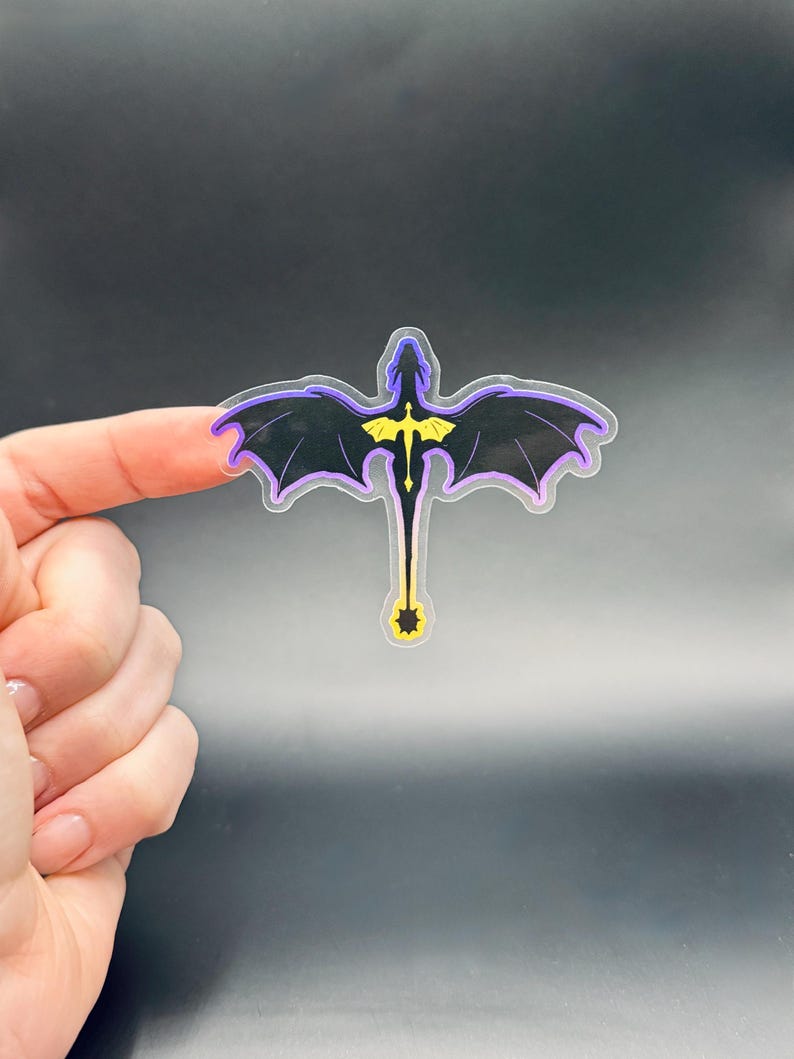 May include: A black dragon sticker with purple and yellow accents. The dragon has outstretched wings and a long tail. The sticker is held between two fingers against a dark background.