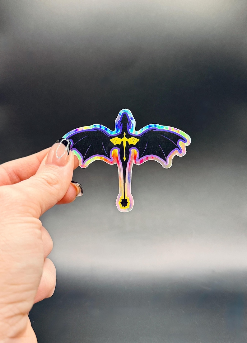 May include: A holographic sticker featuring a stylized dragon with outstretched wings. The dragon is primarily black with iridescent edges and a yellow emblem on its chest. The sticker is held against a dark background.