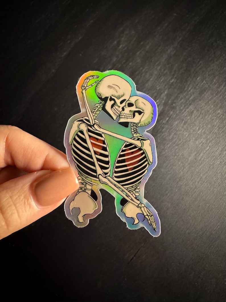 May include: Holographic sticker featuring two skeletons embracing and kissing. The skeletons are depicted in a detailed, anatomical style with visible rib cages and skulls. The sticker has a rainbow iridescent effect, creating a colorful sheen.
