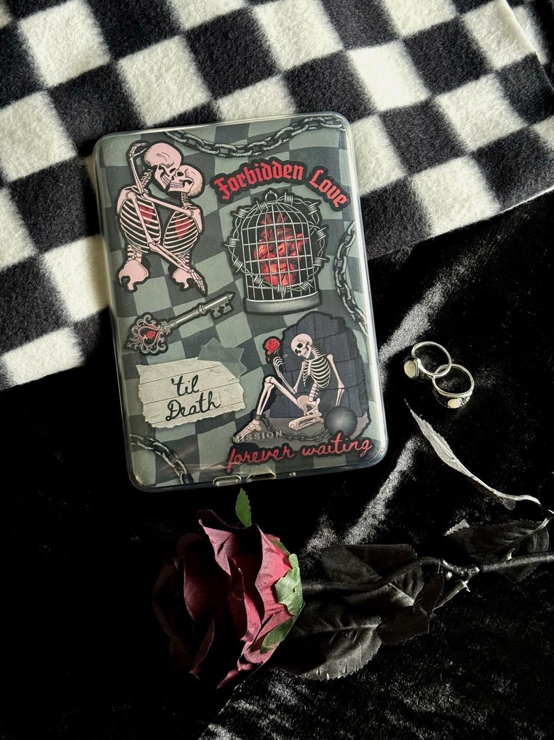 Op de afbeelding: Een doorzichtige telefoonhoes met een gothic thema, met illustraties van skeletten, kettingen en de zinnen "Forbidden Love", "Til Death" en "forever waiting". Een rode roos en zilveren ringen zijn ook zichtbaar.
