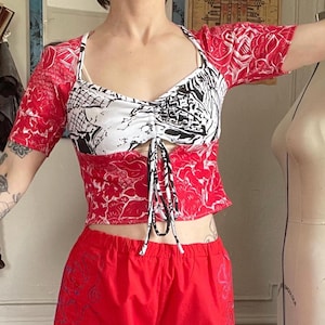 May include: A two-piece outfit featuring a red and white patterned top with short sleeves and a drawstring detail. The top is paired with matching red shorts. The top has a white and black graphic design on the front.