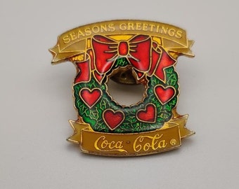 May include: A gold-toned Coca-Cola pin featuring a Christmas wreath design. The wreath is green with red heart accents and a red bow. The pin has a banner that reads "SEASONS GREETINGS" and another with the Coca-Cola logo.