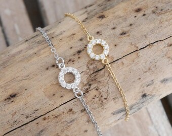 Puede incluir: Dos pulseras delicadas con un colgante circular incrustado con pequeñas piedras transparentes. Una pulsera es plateada y la otra es dorada.