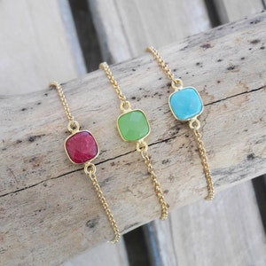 May include: Three gold chain bracelets with square-shaped gemstone charms. The charms are red, green, and blue.