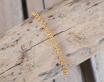 Puede incluir: Una delicada pulsera de tono dorado que presenta una cadena continua de pequeñas cuentas esféricas uniformes unidas en línea recta. La pulsera tiene una superficie pulida y reflectante con un patrón repetitivo constante en toda su longitud.