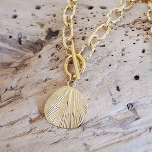 Op de afbeelding: Een goudkleurige ketting met een tuimelsluiting en een hanger in de vorm van een getextureerde zeeschelp. De ketting heeft een ovaal schakelontwerp. De ketting wordt getoond op een stuk drijfhout.