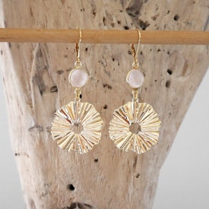 Op de afbeelding: Gouden oorbellen met een delicate roze steen en een getextureerd, zonnestralen-vormig ontwerp.