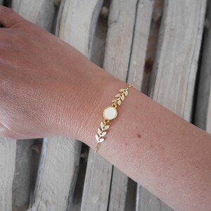 Op de afbeelding: Gouden armband met een witte ronde bedel en een delicaat bladontwerp.