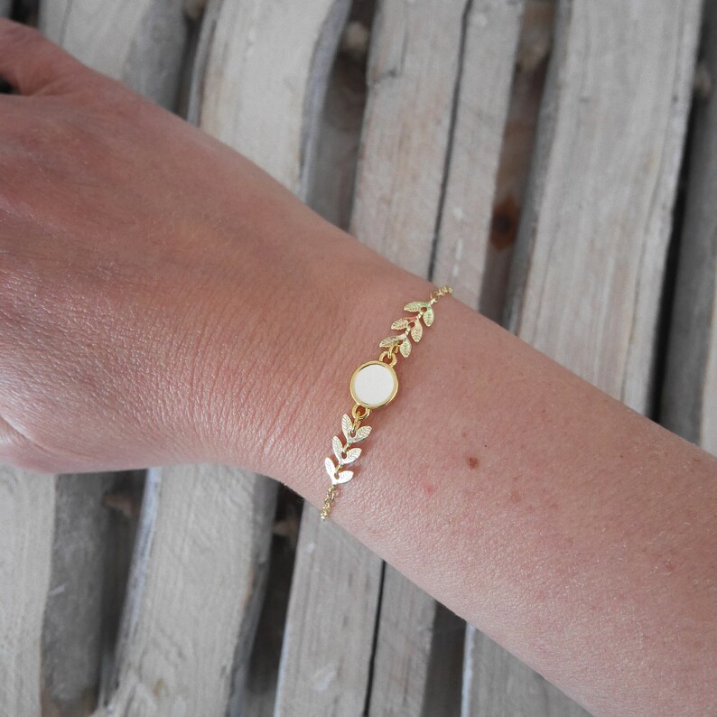 Op de afbeelding: Gouden armband met een witte ronde bedel en een delicaat bladontwerp.