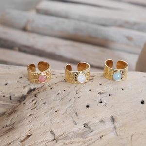 May include: Three gold rings with different colored stones. The rings have a textured design and are set on a wooden surface.