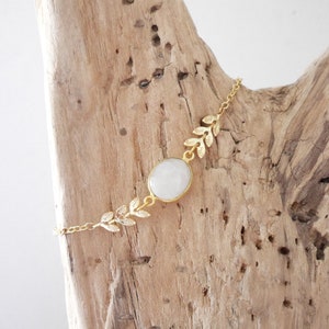 Puede incluir: Un brazalete de cadena dorada con una piedra ovalada blanca y detalles de hojas doradas.