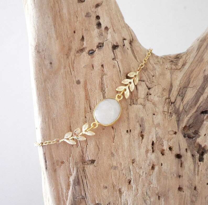 Puede incluir: Un brazalete de cadena dorada con una piedra ovalada blanca y detalles de hojas doradas.