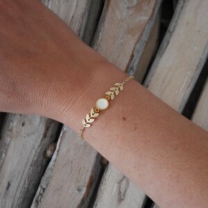 Op de afbeelding: Een gouden kettingarmband met een witte ronde bedel en blaadjesvormige accenten.