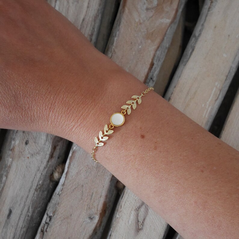 Op de afbeelding: Een gouden kettingarmband met een witte ronde bedel en blaadjesvormige accenten.