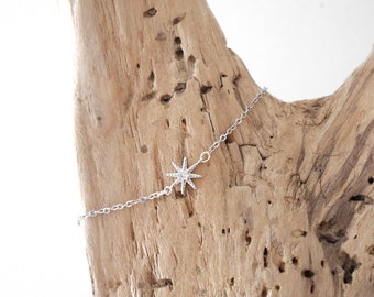 Puede incluir: Una delicada pulsera de cadena en tono plateado con un dije central en forma de estrella de ocho puntas. La estrella está adornada con pequeñas piedras claras y brillantes incrustadas en el metal, creando una superficie brillante y reflectante.