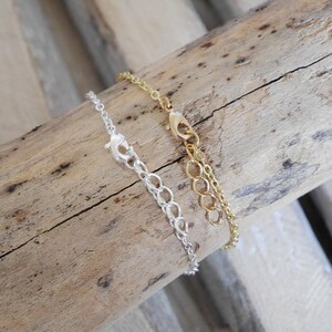 Puede incluir: Dos collares de cadena, uno plateado y otro dorado, con cierres de langosta. Los collares se muestran sobre una superficie de madera.