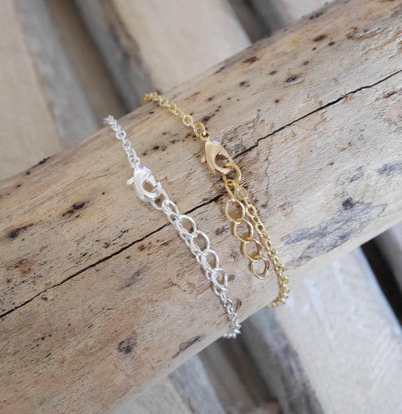 Puede incluir: Dos collares de cadena, uno plateado y otro dorado, con cierres de langosta. Los collares se muestran sobre una superficie de madera.