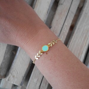 Op de afbeelding: Een gouden armband met een delicaat bladontwerp en een lichtblauwe ronde bedel.