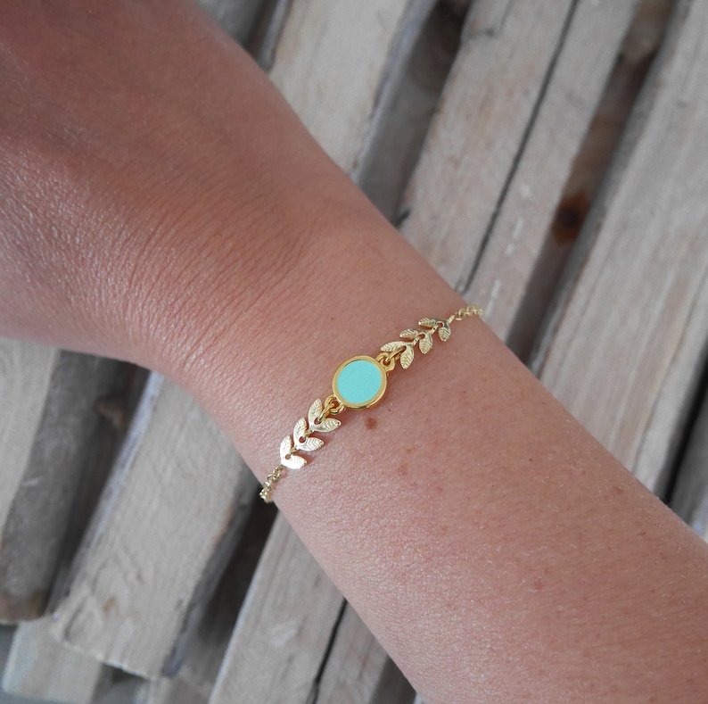 Op de afbeelding: Een gouden armband met een delicaat bladontwerp en een lichtblauwe ronde bedel.