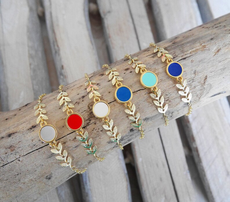 Op de afbeelding: Vijf gouden bladeren armbanden met verschillende gekleurde emaille cirkels. De cirkels zijn rood, wit, turquoise, donkerblauw en lichtblauw.