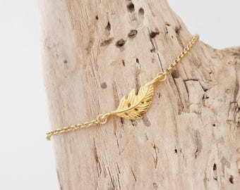 Puede incluir: Un delicado brazalete de cadena dorada con un solo dije de pluma detallado. El brazalete se muestra sobre una superficie de madera marrón claro.