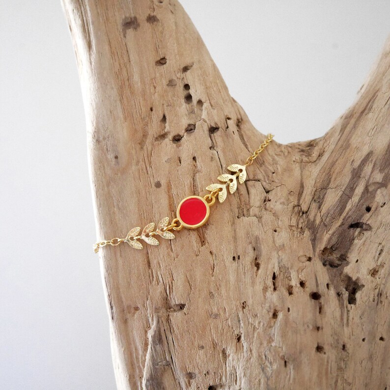 Op de afbeelding: Een gouden kettingarmband met een rode ronde hanger en blaadjesvormige bedels.