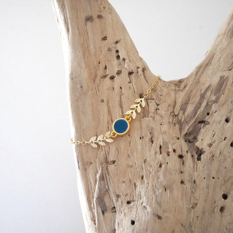 Op de afbeelding: Een gouden kettingarmband met een blauwe ronde bedel en gouden bladeren accenten.