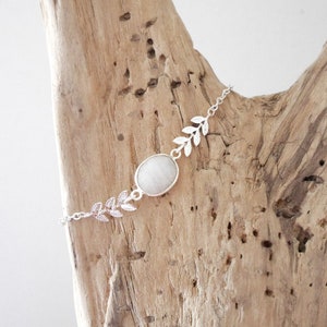 Puede incluir: Un brazalete de cadena de plata con una piedra ovalada blanca y detalles de hojas.