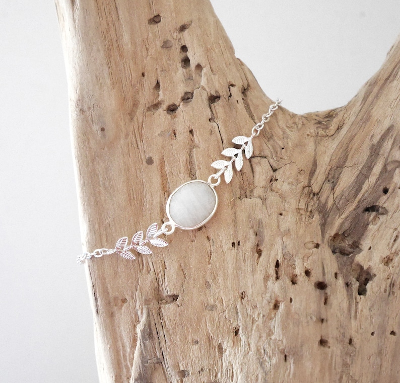 Puede incluir: Un brazalete de cadena de plata con una piedra ovalada blanca y detalles de hojas.
