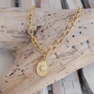 Op de afbeelding: Een gouden ketting met een getextureerde ovale hanger met een klein, helder edelsteentje in het midden. De ketting wordt getoond op een stuk drijfhout.