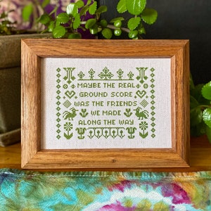 May include: A framed cross-stitch with green embroidery on white fabric. The text reads "Maybe the real ground score was the friends we made along the way."
