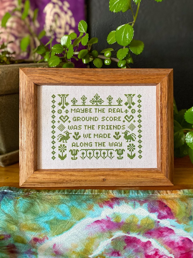 May include: A framed cross-stitch with green embroidery on white fabric. The text reads "Maybe the real ground score was the friends we made along the way."