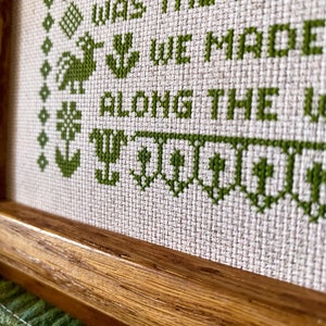 May include: A framed cross-stitch with green embroidery thread on a beige fabric. The text reads "We grew up, we were the friends we made along the way".