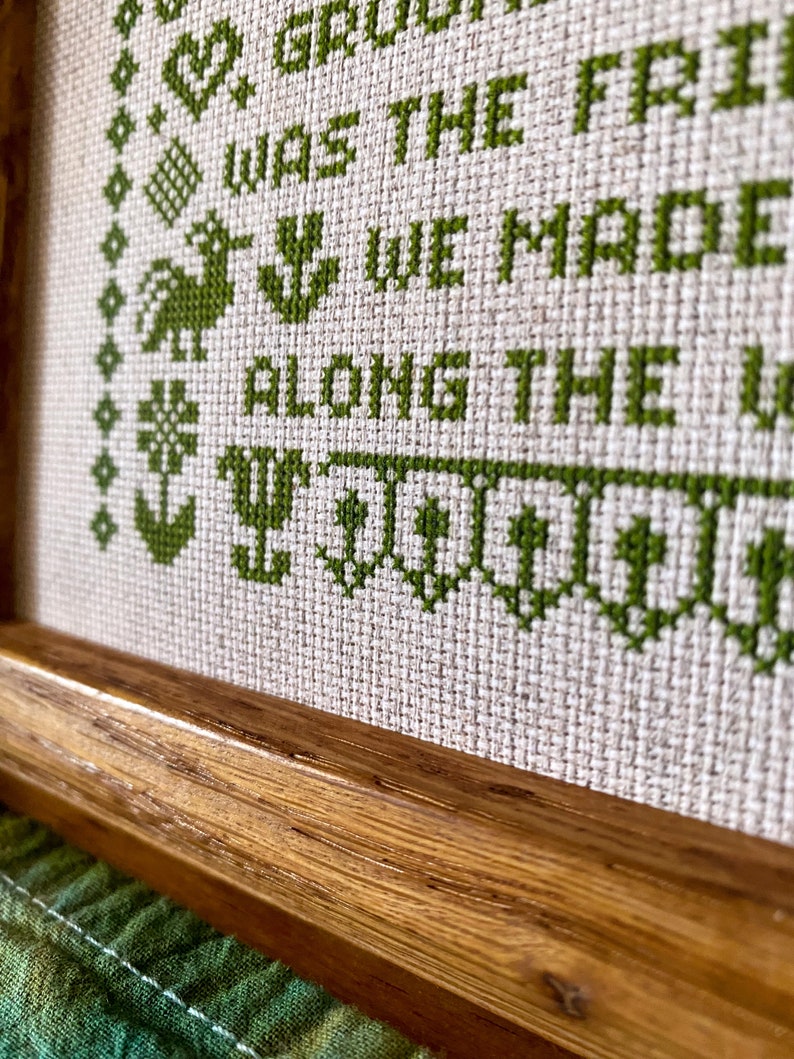 May include: A framed cross-stitch with green embroidery thread on a beige fabric. The text reads "We grew up, we were the friends we made along the way".