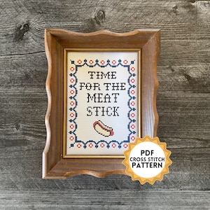 May include: A framed cross stitch pattern with the text "TIME FOR THE MEAT STICK" and a small image of a hot dog. The frame is brown wood with a wavy edge. A yellow star sticker with the text "PDF CROSS STITCH PATTERN" is below the frame.