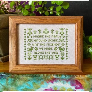 May include: A framed cross-stitch embroidery with green thread on white fabric. The embroidery reads "Maybe the real ground score was the friends we made along the way".